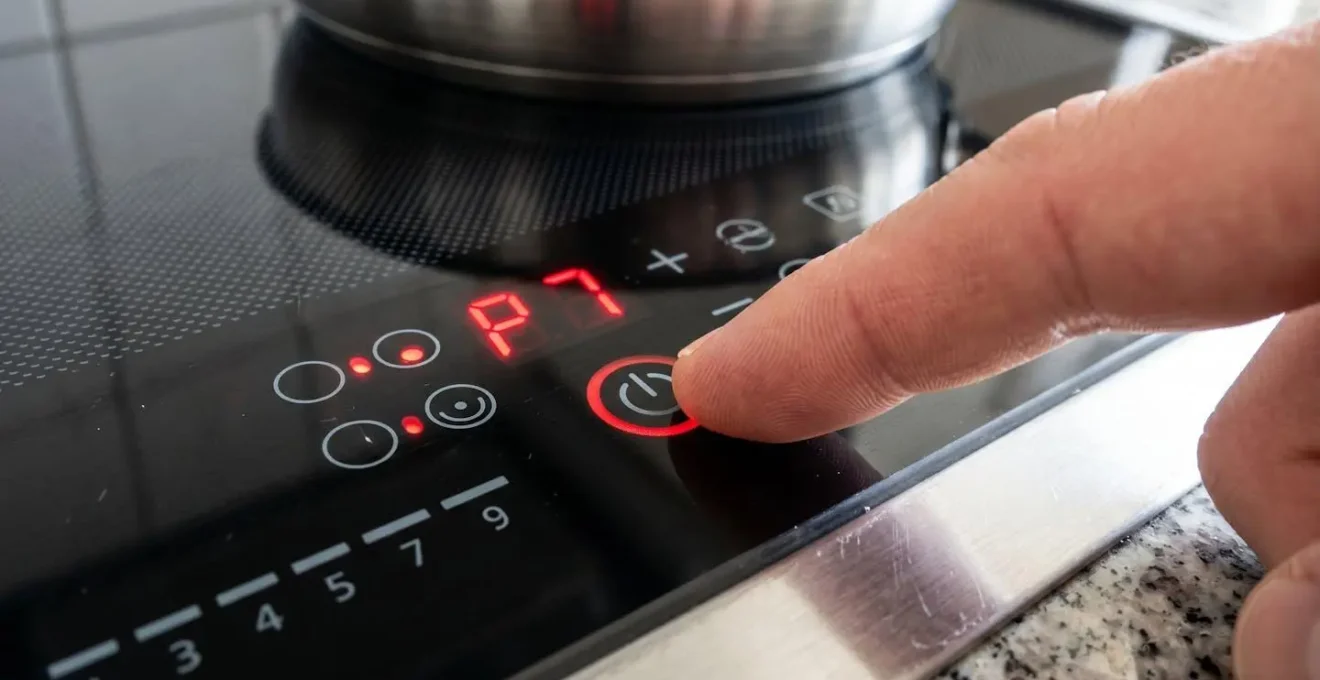 Gros plan serré sur les commandes tactiles d'une plaque à induction, doigt effleurant le symbole de puissance, affichage LED rouge indiquant le niveau sept
